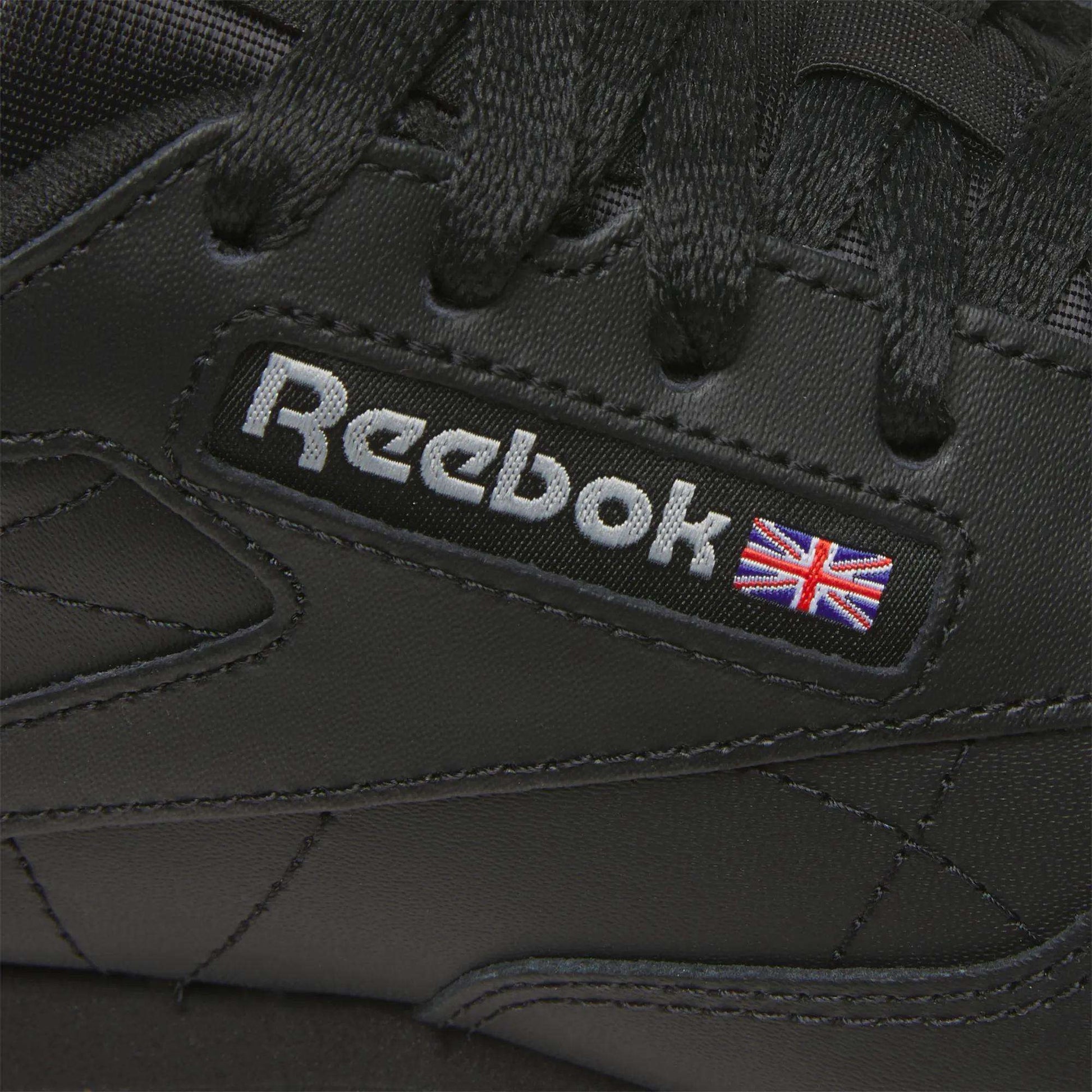 Close-up of Reebok Women's Classic Leather Shoes in black, showcasing the Reebok logo, a small Union Jack flag by the laces, and a cushioned midsole for comfort.