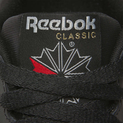 Close-up of a Women's Reebok Classic Leather Shoe tongue, showing the white Reebok logo and CLASSIC in gold above a stylized maple leaf, with black laces in front and a cushioned midsole for comfort.