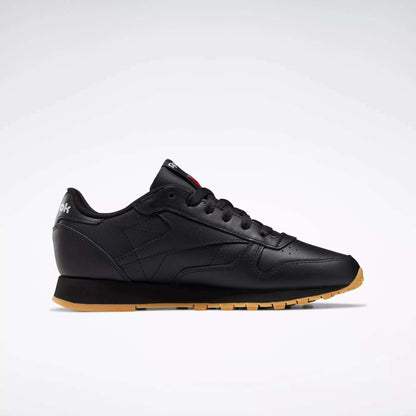 The Reebok Women's Classic Leather Shoes are shown in side profile with a black leather upper, gum sole, black laces, subtle stitching, and a cushioned midsole for comfort, set against a plain white background.