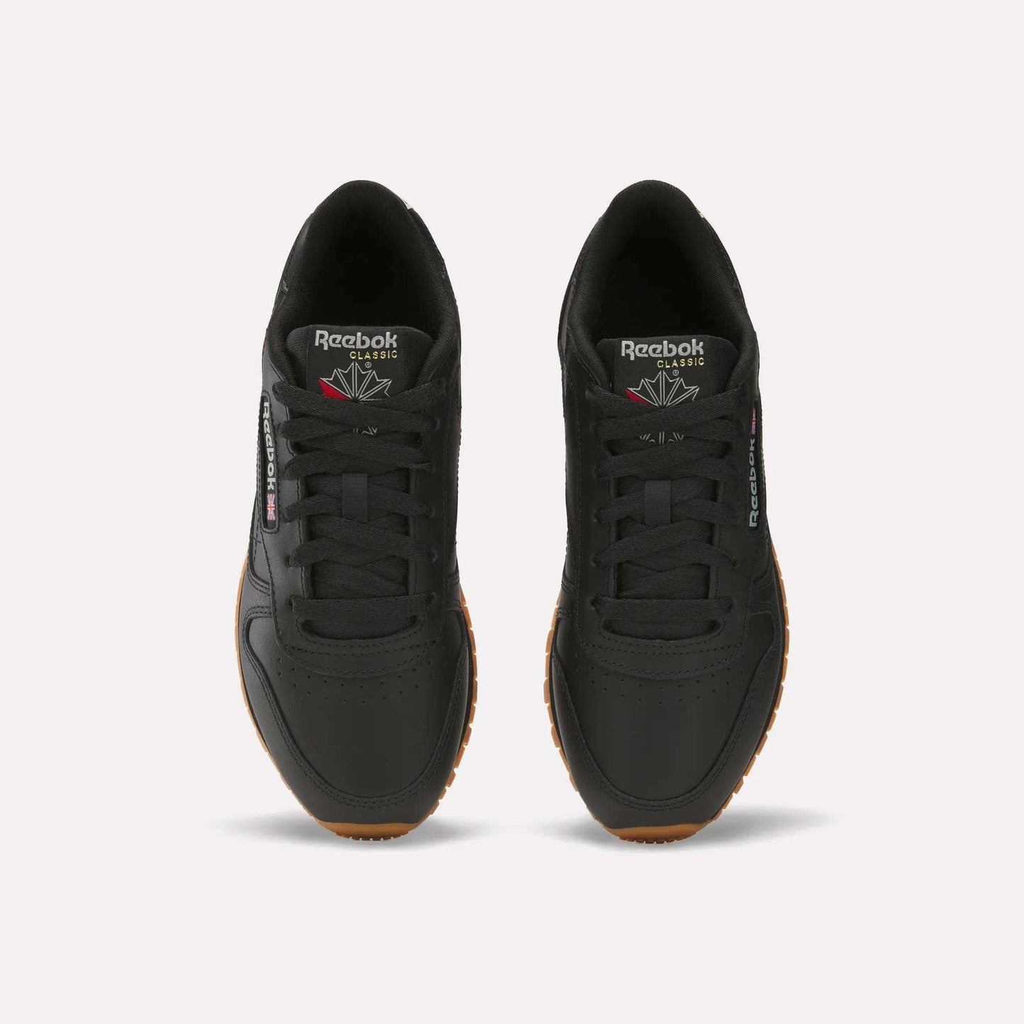 A pair of Women's Classic Leather Shoes by Reebok in black with gum soles, shown from above. They display the Reebok logo on the tongue and side, feature cushioned midsoles, and have neatly tied laces for comfort and style.