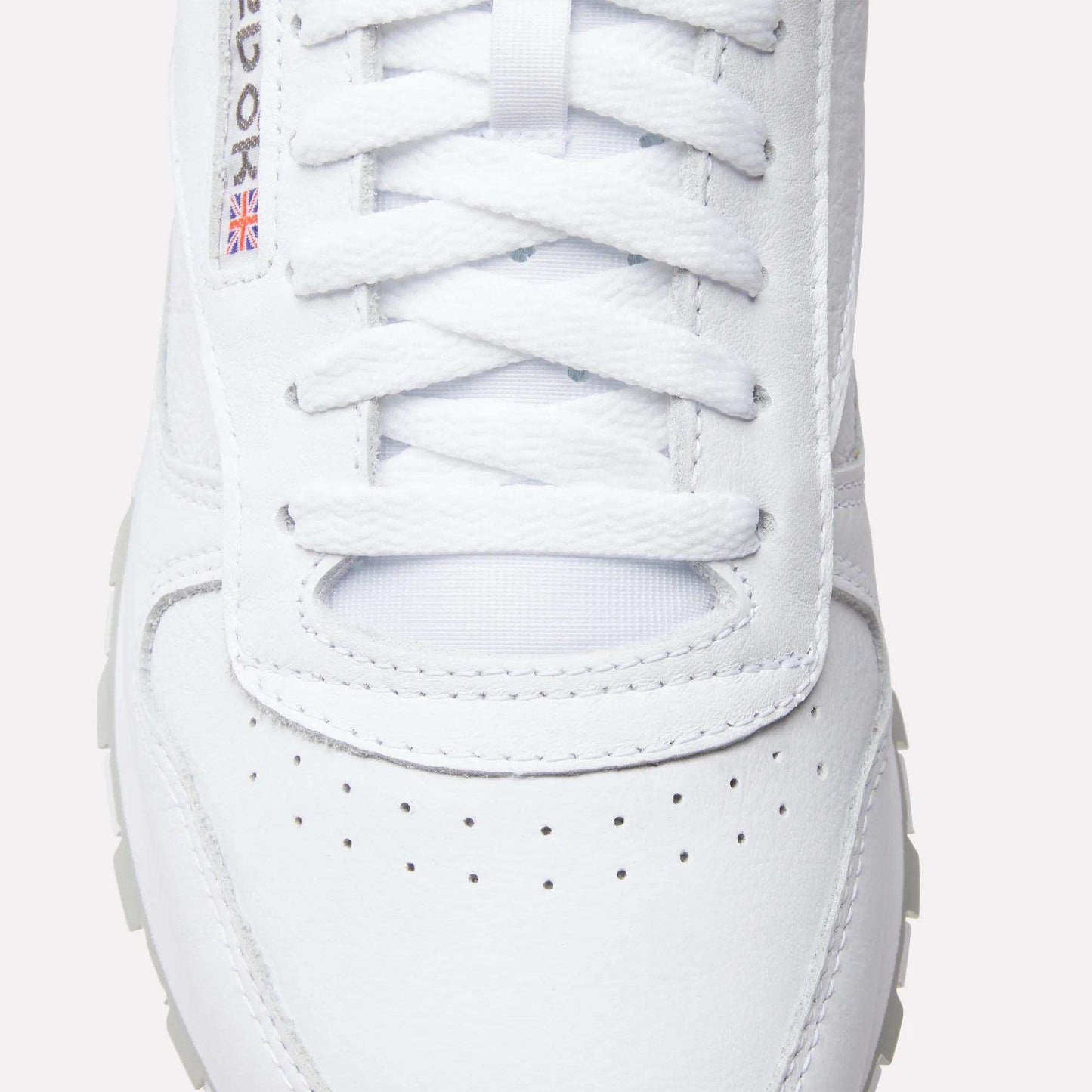 Close-up of Reebok Classic Leather Shoes with laces, a perforated toe box, textured leather, and a small Union Jack flag detail by the eyelets—iconic elements from Reebok’s retro design.