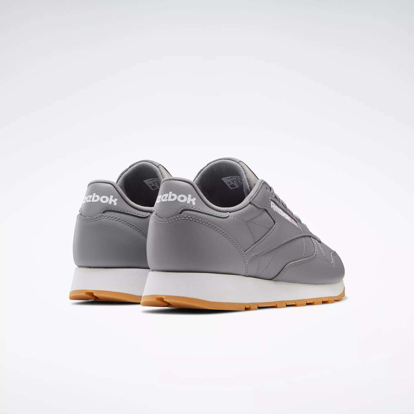 A pair of Reebok Classic Leather Shoes, featuring a gray leather upper, white soles, light brown outsoles, and retro styling with the Reebok logo on the heel, are positioned side by side facing away on a simple light background.