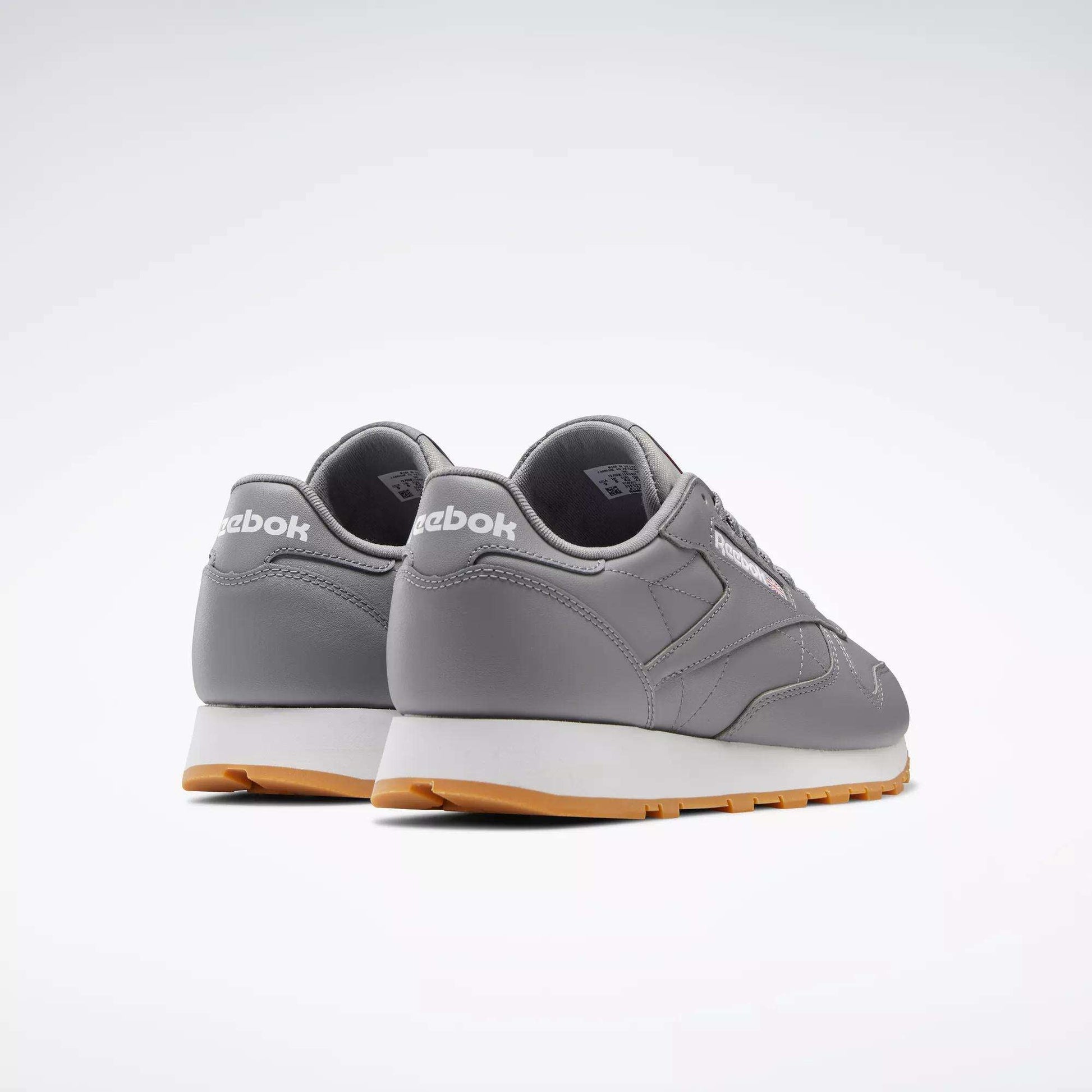 A pair of Reebok Classic Leather Shoes, featuring a gray leather upper, white soles, light brown outsoles, and retro styling with the Reebok logo on the heel, are positioned side by side facing away on a simple light background.