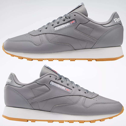 The Reebok Classic Leather Shoes feature gray leather uppers, white midsoles, and light brown rubber outsoles. Seen from the side against a plain background, they display the Reebok logo and UK flag on the sides.