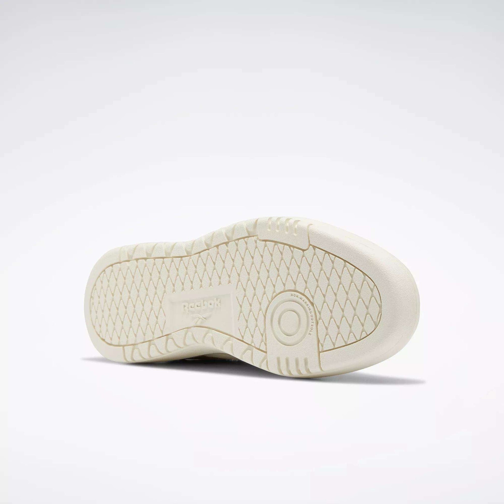 The image displays the sole of a beige Reebok Women's Club C Double Revenge Shoe, featuring a textured, diamond-patterned tread and circular details near the heel, reflecting its court-inspired design and double outsole for enhanced grip.