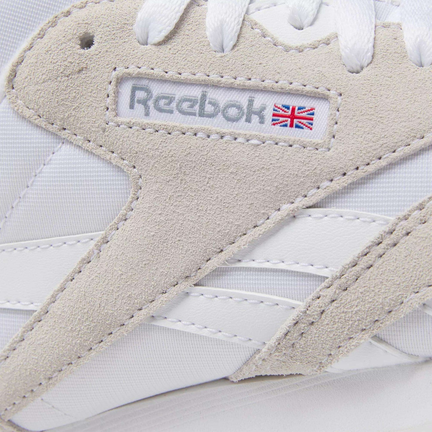 Close-up of Reebok Women's Classic Nylon Shoes in white with beige suede accents, white laces, and the Reebok logo featuring a small UK flag—stylish everyday sneakers.