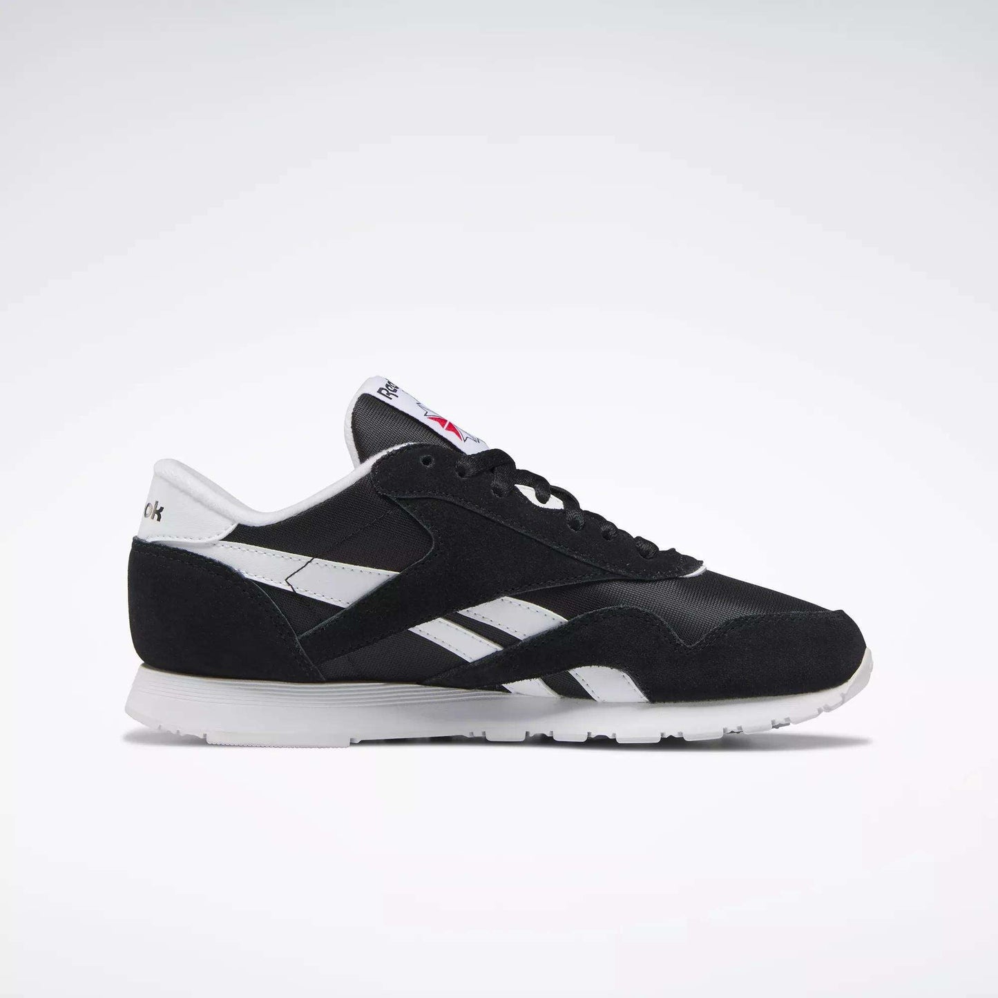 The Reebok Women's Classic Nylon Shoes feature a black upper, white side stripes, suede accents, and a white sole with logo details—shown in side profile on a light background. Stylish and comfortable sneakers for everyday wear.