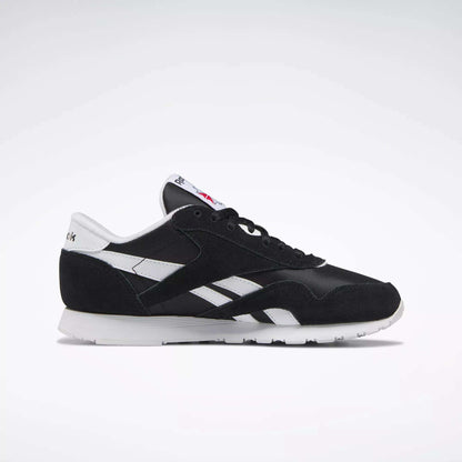 The Reebok Women's Classic Nylon Shoes feature a black upper, white side stripes, suede accents, and a white sole with logo details—shown in side profile on a light background. Stylish and comfortable sneakers for everyday wear.