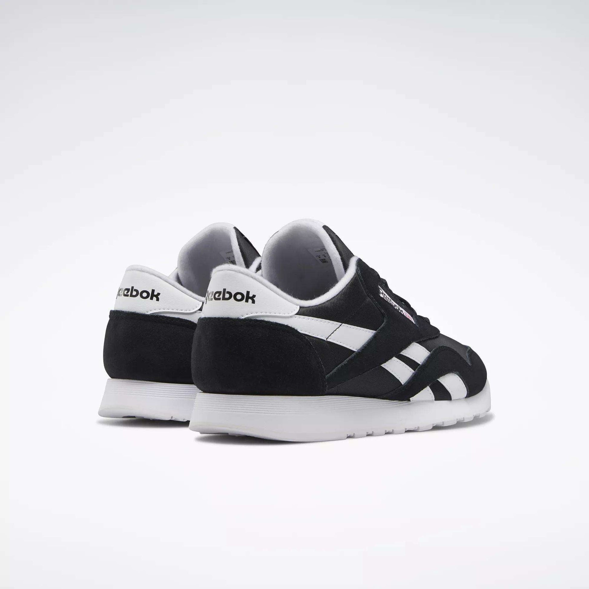 A pair of Men's Classic Nylon Shoes by Reebok in black and white, set on a white background with one shoe behind the other, showcasing side and back views—classic everyday sneakers with timeless style.