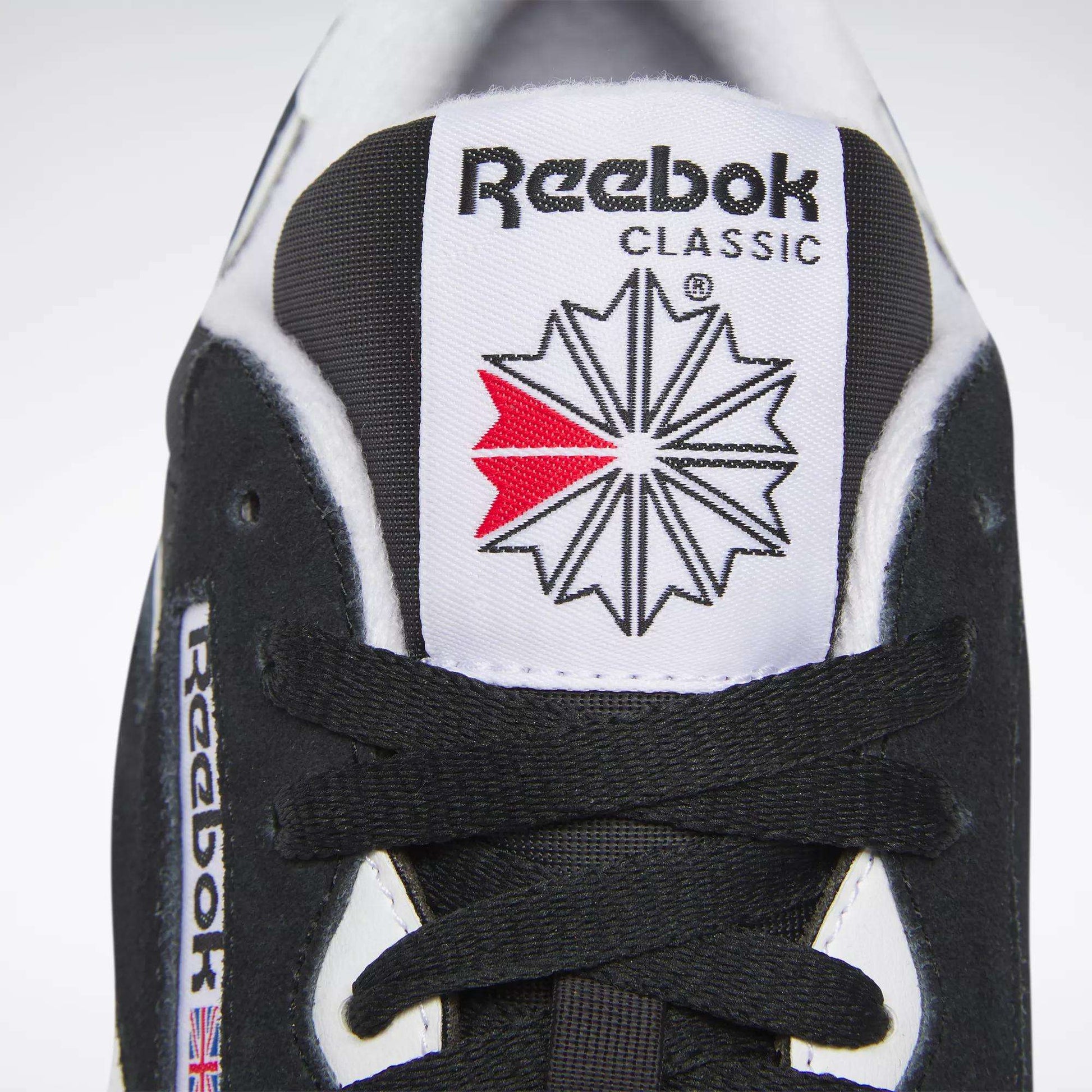 Close-up of the tongue of a black Reebok Men's Classic Nylon Shoe, highlighting the Reebok logo, red and grey starburst, black laces, and a small Union Jack flag—signature details of these classic sneakers.
