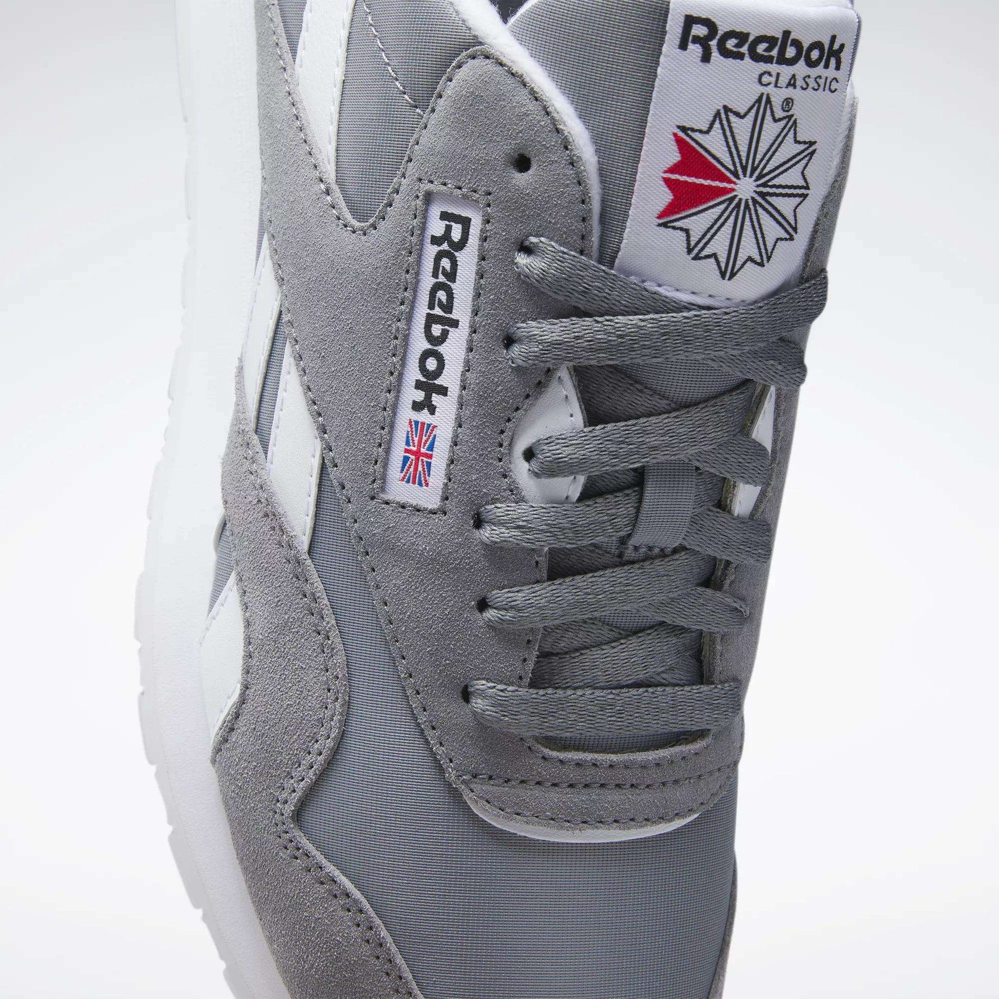 A close-up of Reebok Men's Classic Nylon Shoes with a grey nylon upper, white stripes, laces, Reebok branding, and a red, white, and blue geometric design on the tongue for classic everyday style.