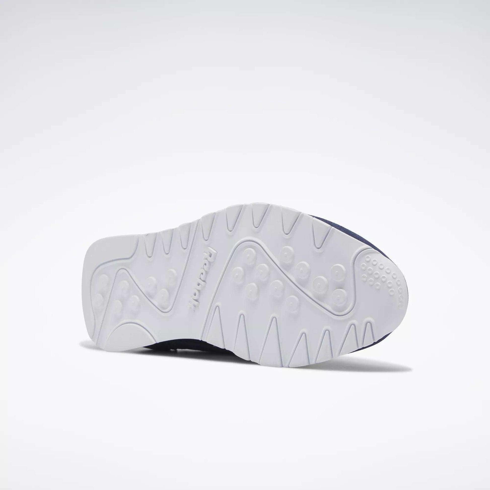 The Reebok Men's Classic Nylon Shoes are shown on their side against a plain light background, displaying the white textured sole with tread patterns and Reebok branding, emphasizing the grip of these classic everyday sneakers.