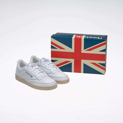 A pair of Women's Club C 85 Vintage Shoes by Reebok, in white with gum soles, are placed next to a shoebox featuring a large Union Jack flag against a plain background.