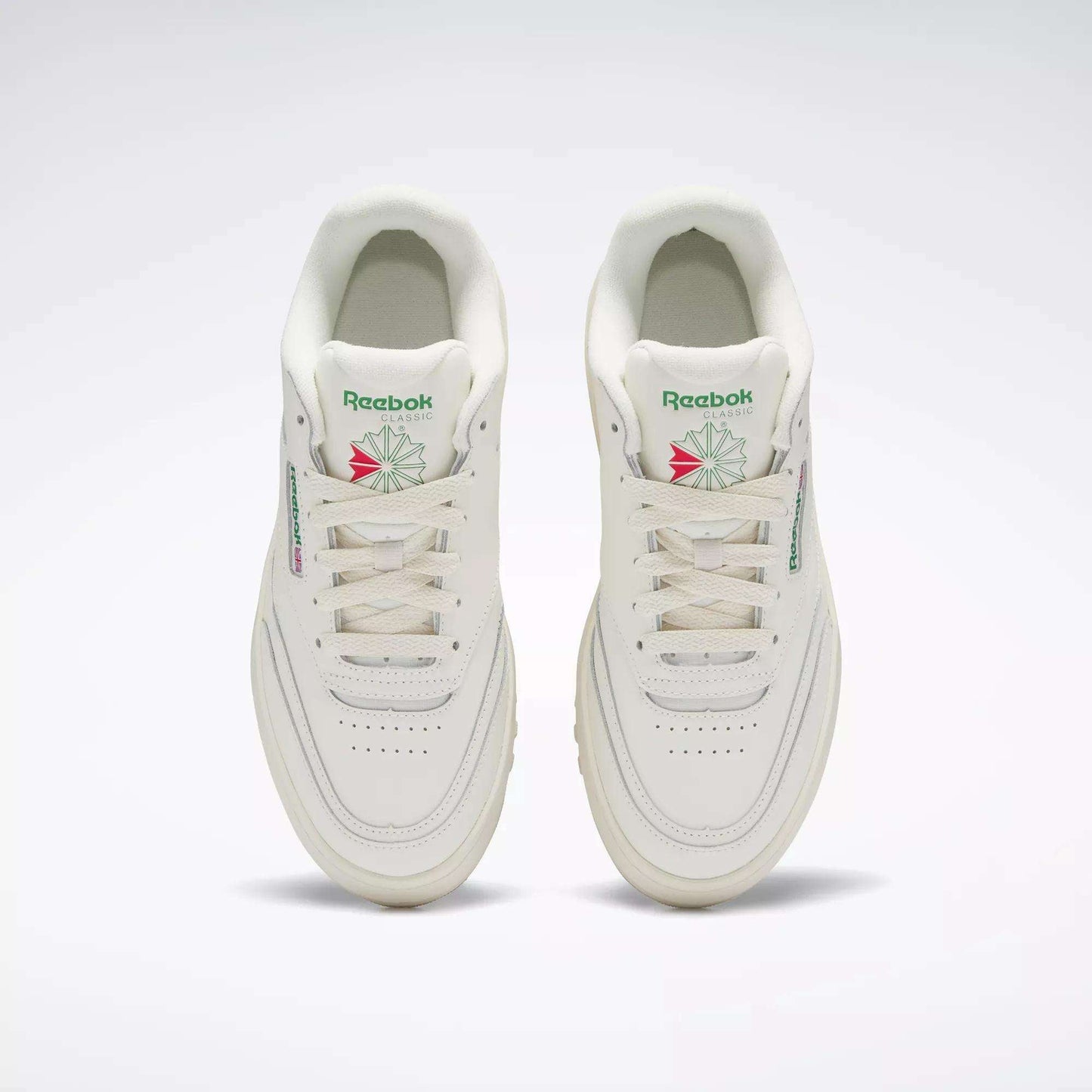 A pair of Reebok Women's Club C Extra Shoes in white, featuring green and pink logo accents and platform midsoles, are shown side by side from above on a plain light background, highlighting the tops, tongues, and laces.