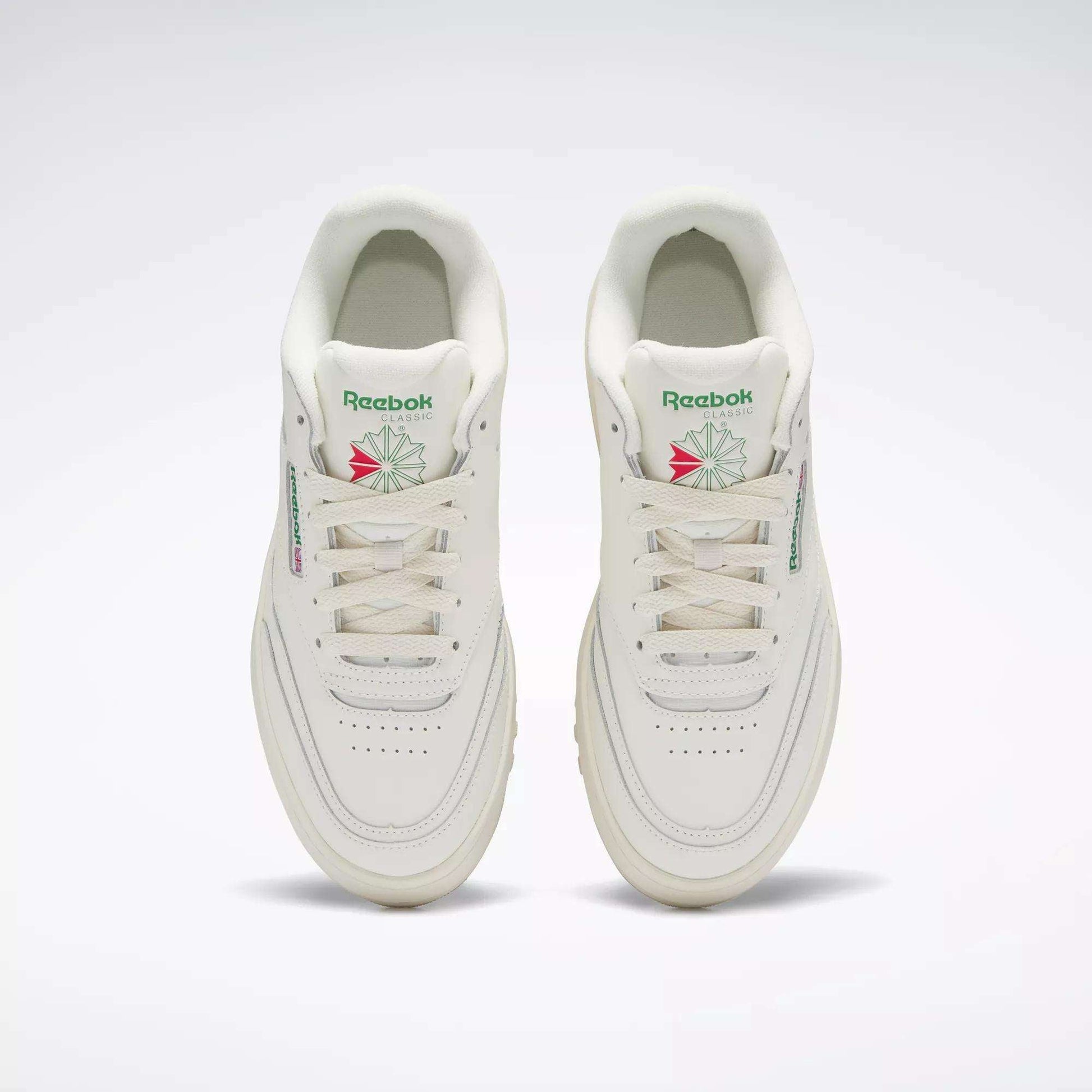 A pair of Reebok Women's Club C Extra Shoes in white, featuring green and pink logo accents and platform midsoles, are shown side by side from above on a plain light background, highlighting the tops, tongues, and laces.