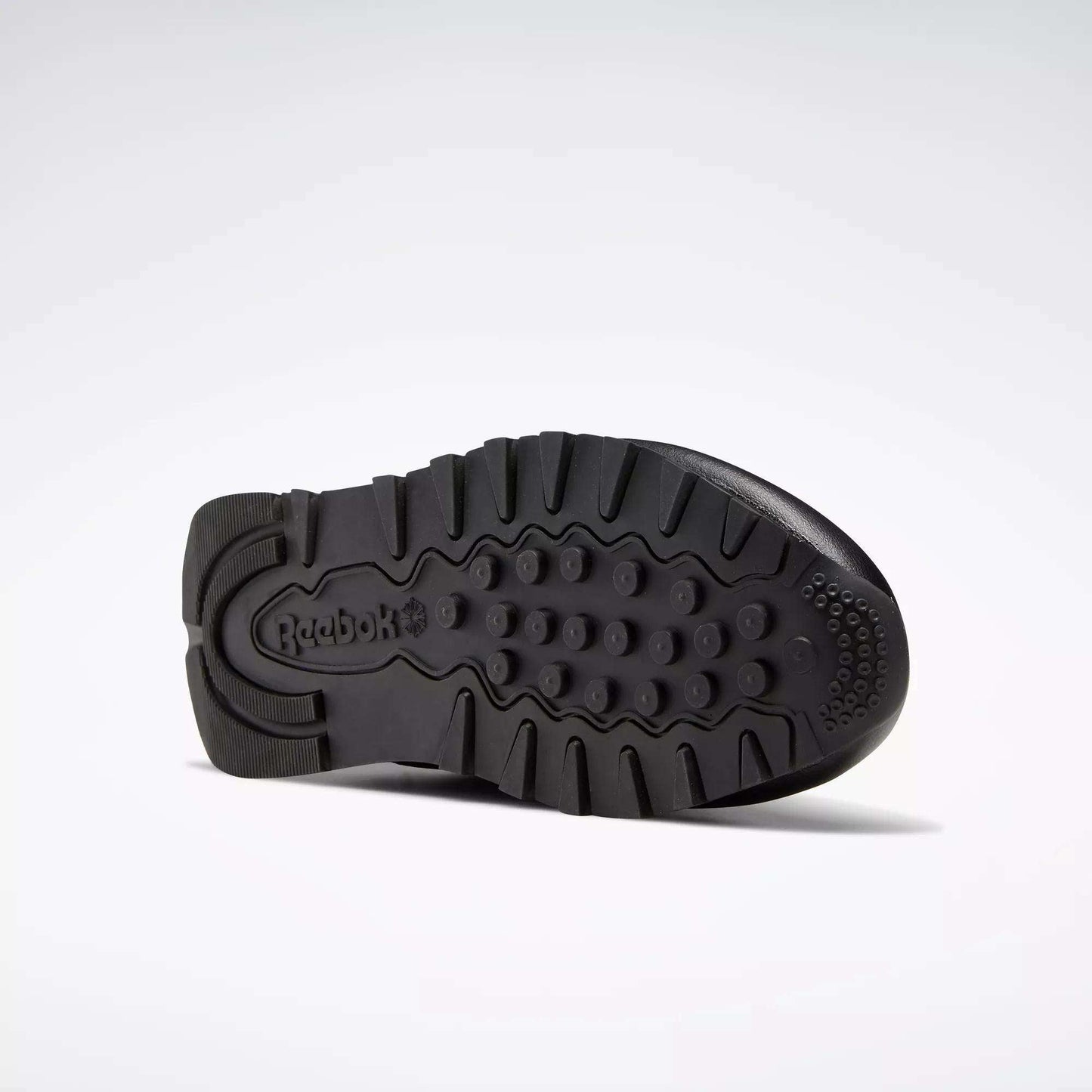 The sole of these Classic Leather Shoes - Big Kids by Reebok, shown in black from the bottom, has textured grip patterns and an embossed Reebok logo near the heel, emphasizing a timeless design.