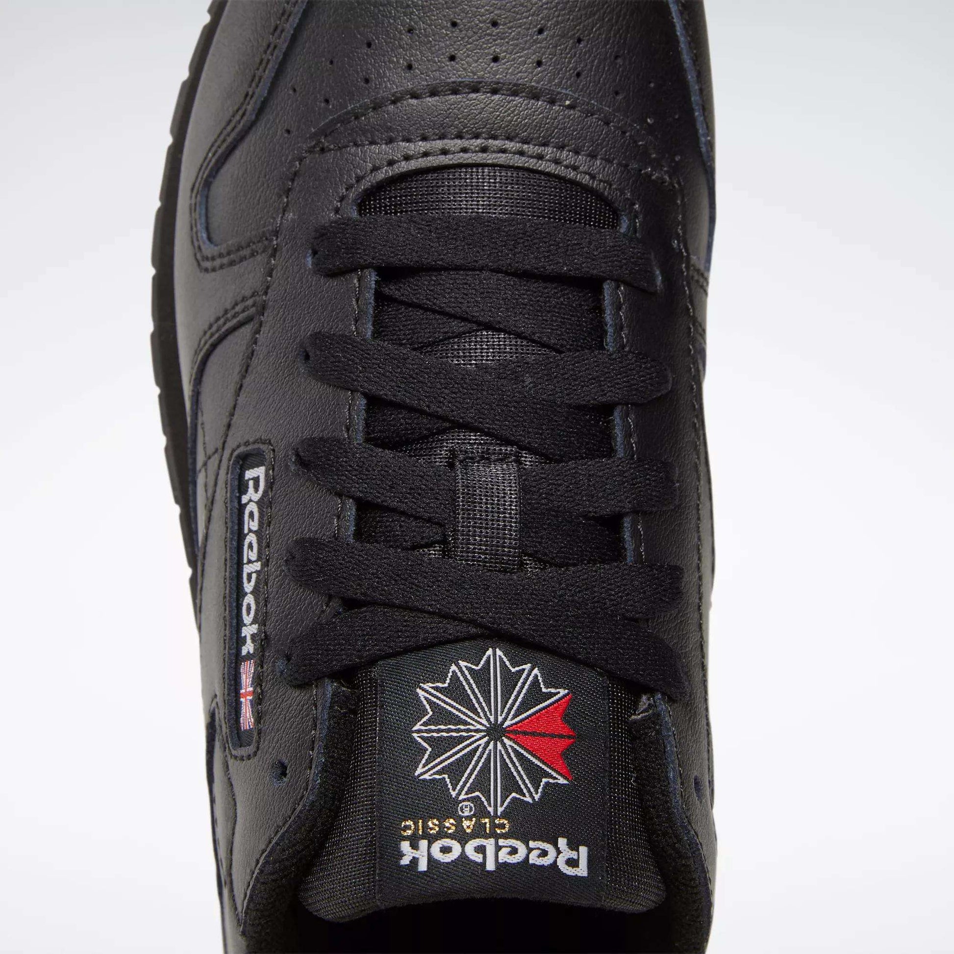 Close-up, top-down view of black Reebok Classic Leather Shoes for big kids, featuring black laces, perforated toe box, and the iconic Union Jack and starburst Reebok logo on the tongue—ideal for boys’ shoe collections.