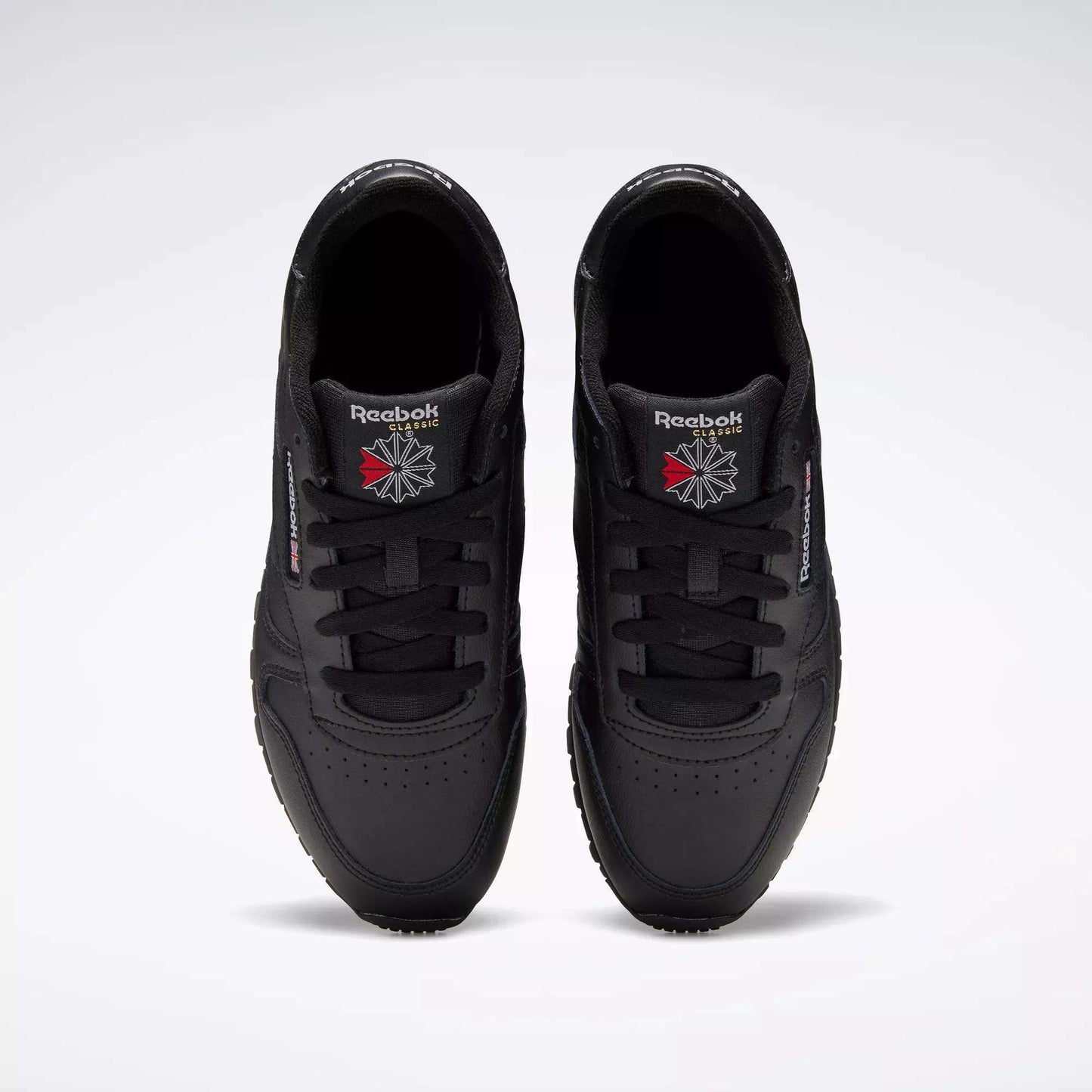 A pair of Reebok Classic Leather Shoes - Big Kids is seen from above, displaying the black uppers and logos on the tongues against a plain light gray background.