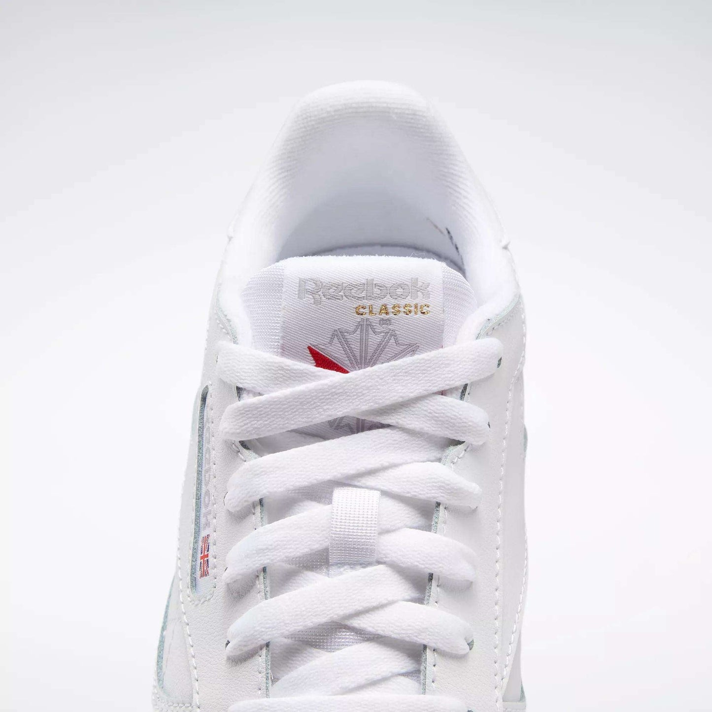 A close-up of the Reebok Classic Leather Shoes - Big Kids highlights their timeless white design, showing the laces, tongue, and a gold Reebok logo with a red emblem above it.