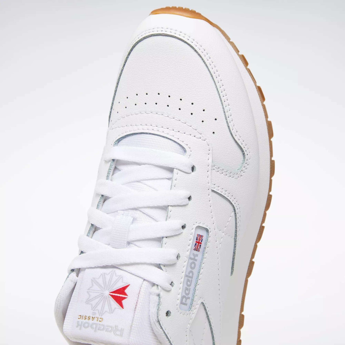 A close-up of the Reebok Classic Leather Shoes - Big Kids in white, featuring a gum sole, logo, red and gray accents, stitched side-stripe, perforated toe box, and white laces.