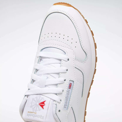 A close-up of the Reebok Classic Leather Shoes - Big Kids in white, featuring a gum sole, logo, red and gray accents, stitched side-stripe, perforated toe box, and white laces.