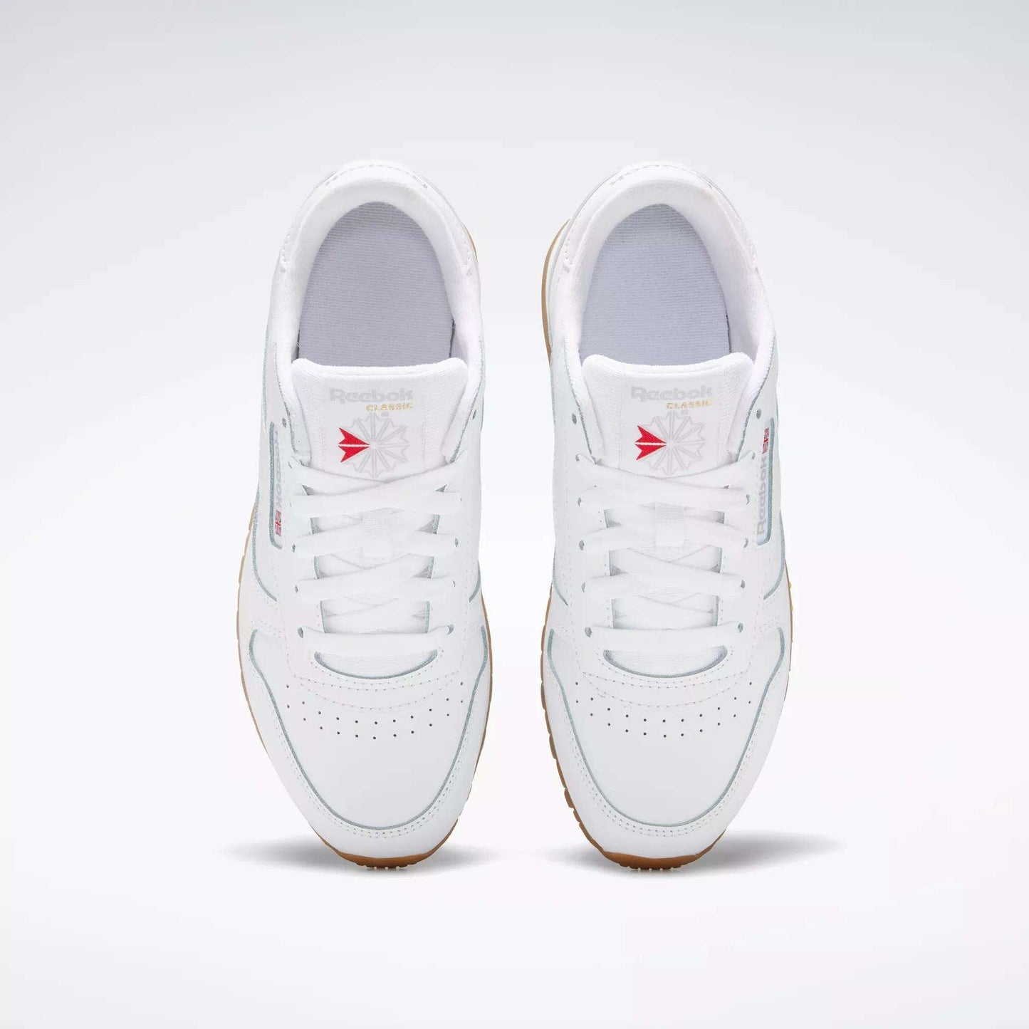 A pair of white Reebok Classic Leather Shoes for big kids, with gum soles and stitched side stripes, viewed from above. Red accents on the tongue stand out as the shoes are positioned side by side on a light gray background.