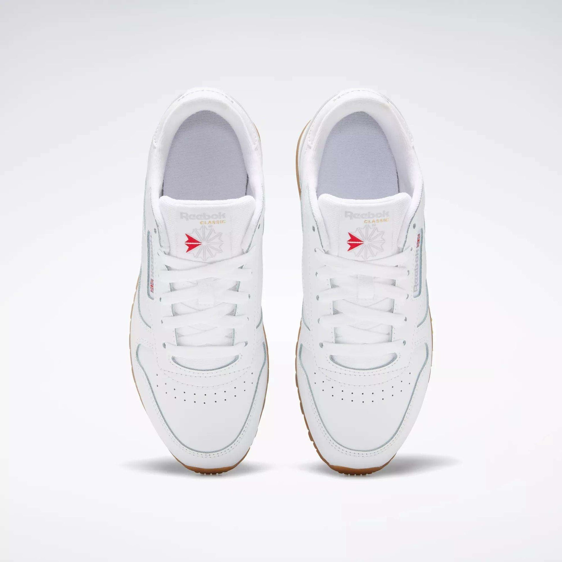 A pair of white Reebok Classic Leather Shoes for big kids, with gum soles and stitched side stripes, viewed from above. Red accents on the tongue stand out as the shoes are positioned side by side on a light gray background.