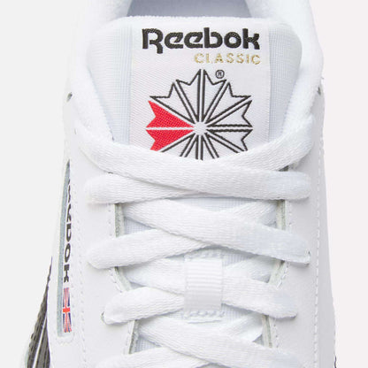 Close-up of a white Reebok Club C Revenge shoe displaying the tongue with the Reebok logo, red and black star design, and white laces. The Reebok name and small UK flag are visible on these casual leather shoes.