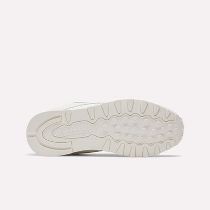The image shows the bottom of a white Reebok Classic Leather Shoe, featuring a textured sole for grip and the Reebok logo near the heel, emphasizing its durable build and retro style. Product: Classic Leather Shoes by Reebok.