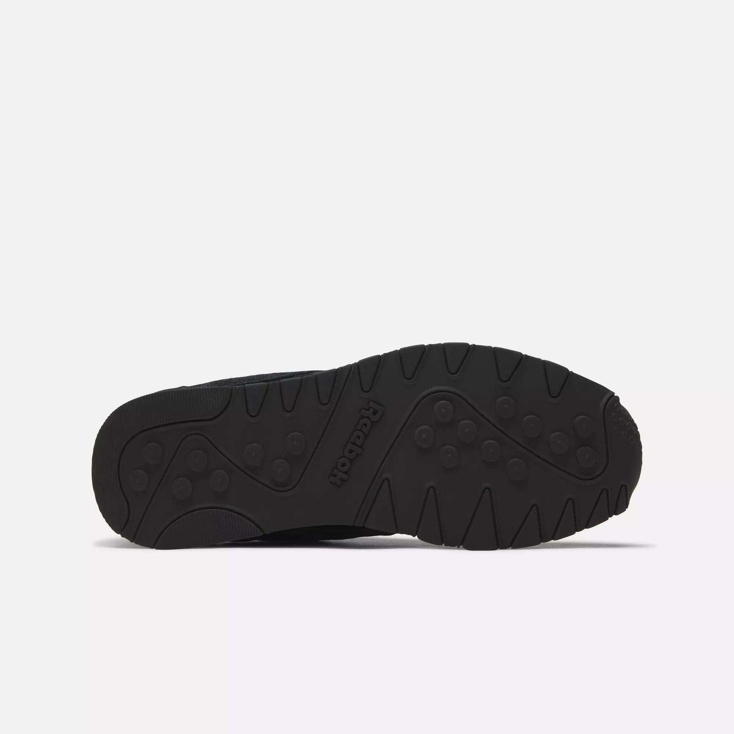 The image shows the black rubber outsole of Reebok Men's Classic Nylon Shoes, featuring a textured tread with circular and linear grooves for grip. The Reebok logo is embossed in the center of the sole.