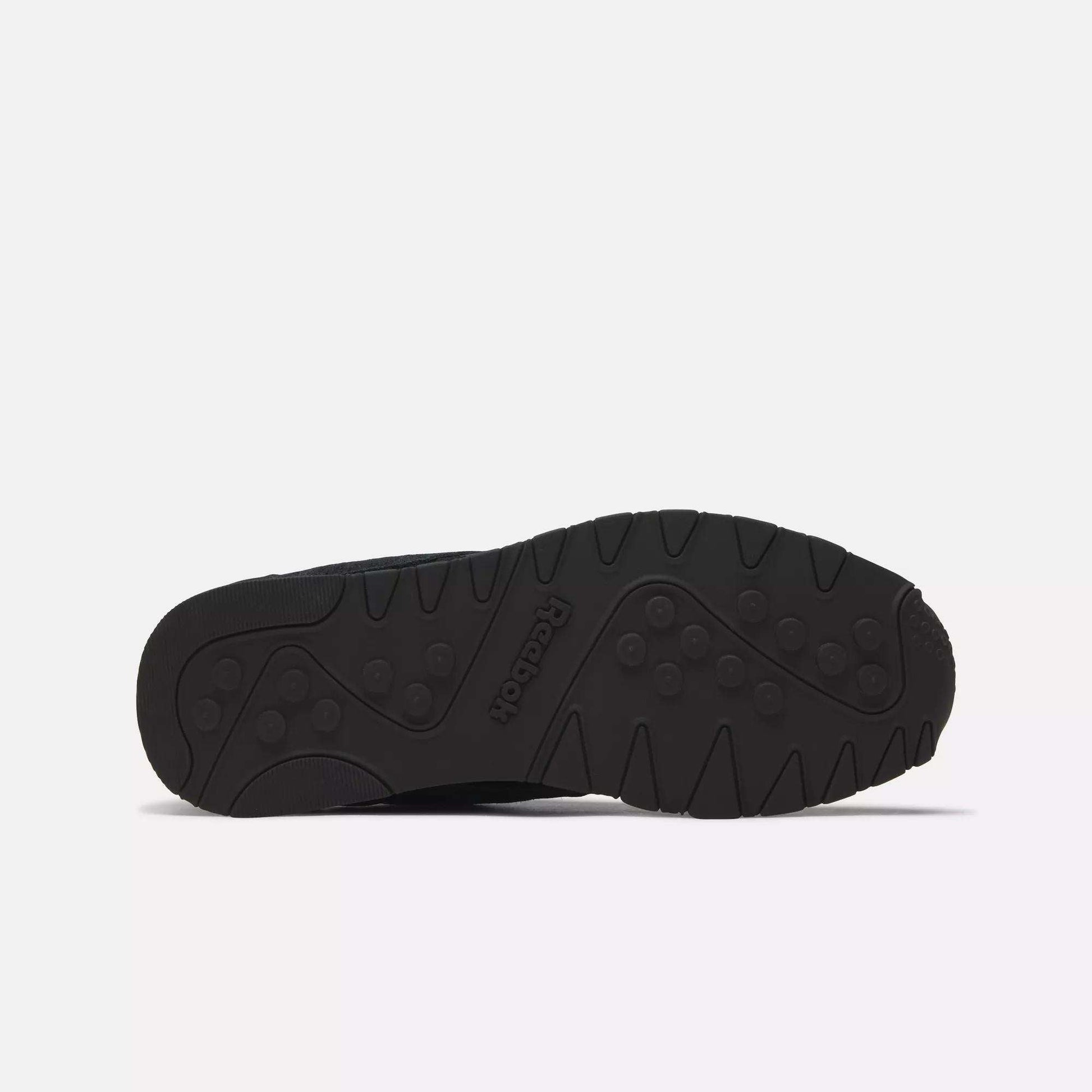 The image shows the black rubber outsole of Reebok Men's Classic Nylon Shoes, featuring a textured tread with circular and linear grooves for grip. The Reebok logo is embossed in the center of the sole.