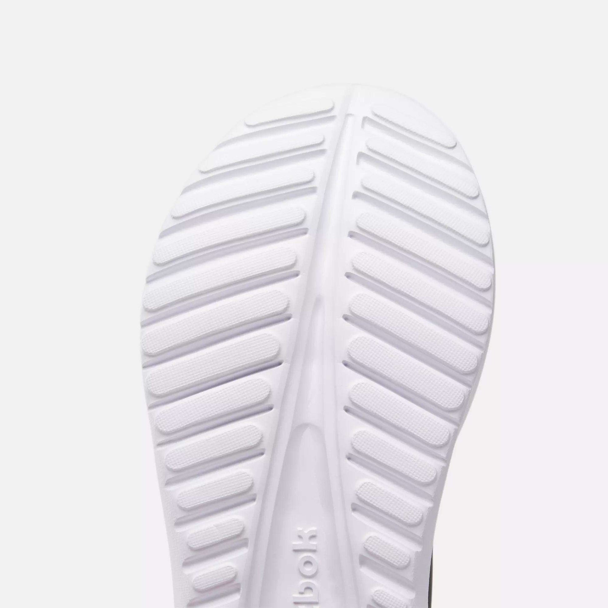 Close-up of the sole of a white Reebok Women's Energen Lux Running Shoe, featuring horizontal and diagonal grooves for traction. The lightweight, ribbed sole has a textured pattern with the Reebok logo partially visible.
