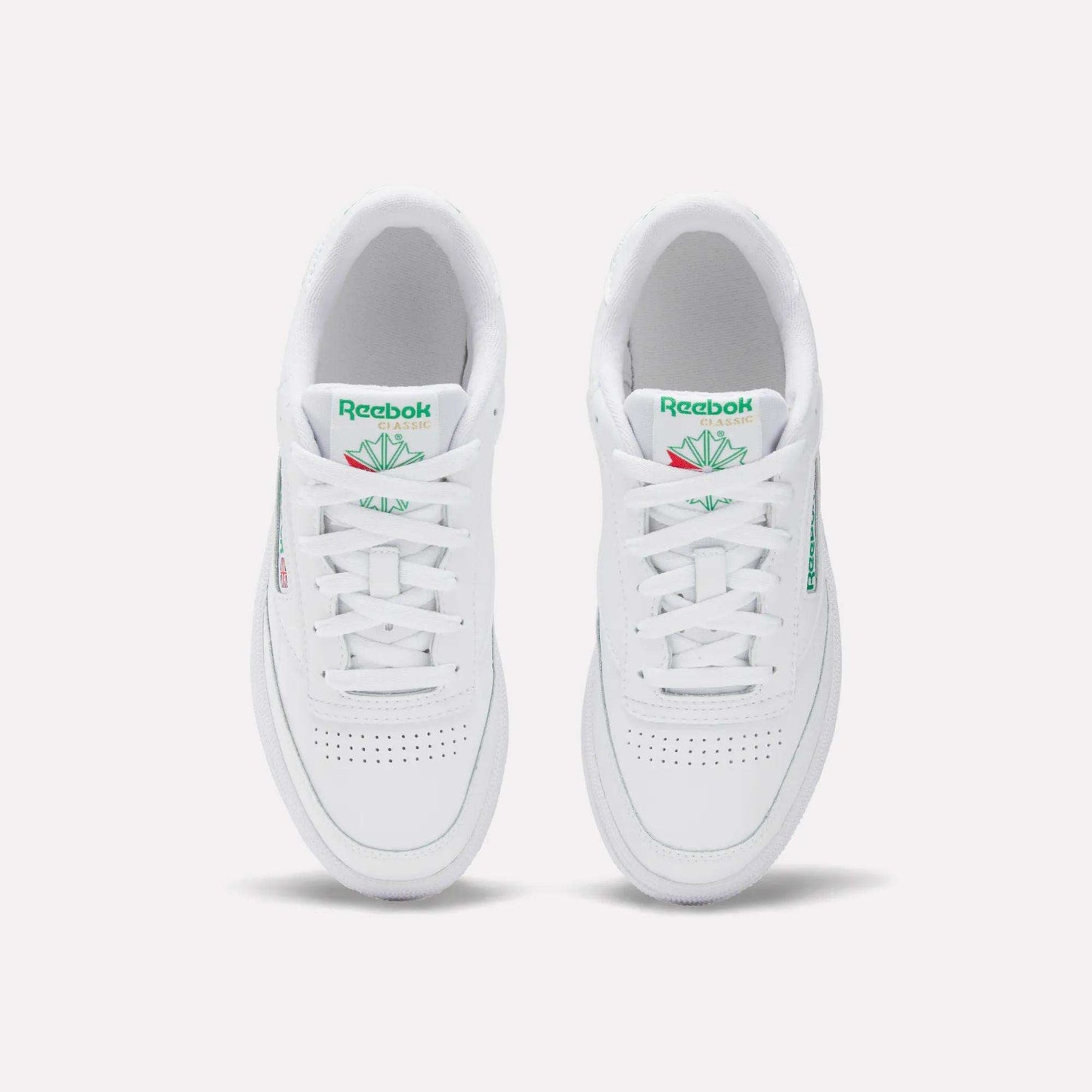 The Women's Club C 85 Shoes by Reebok are white sneakers with green Reebok logos and tongue detailing, a clean minimalist look, and toe perforations, shown from an overhead view.