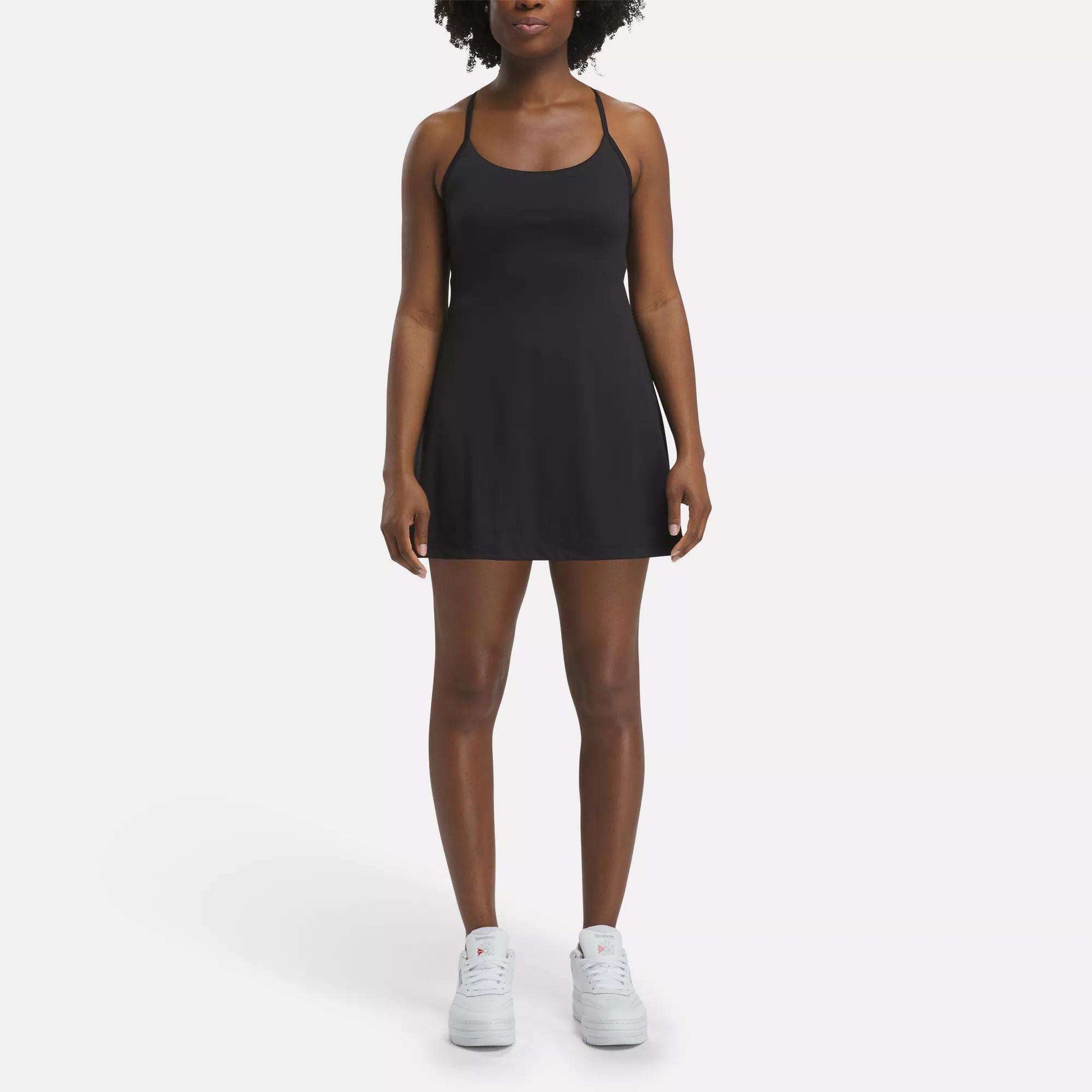 A woman faces forward against a plain white background, wearing the Reebok Women's Lux Strappy Dress in black with white sneakers.