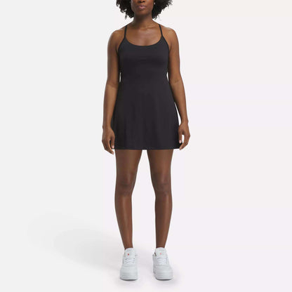 A woman faces forward against a plain white background, wearing the Reebok Women's Lux Strappy Dress in black with white sneakers.