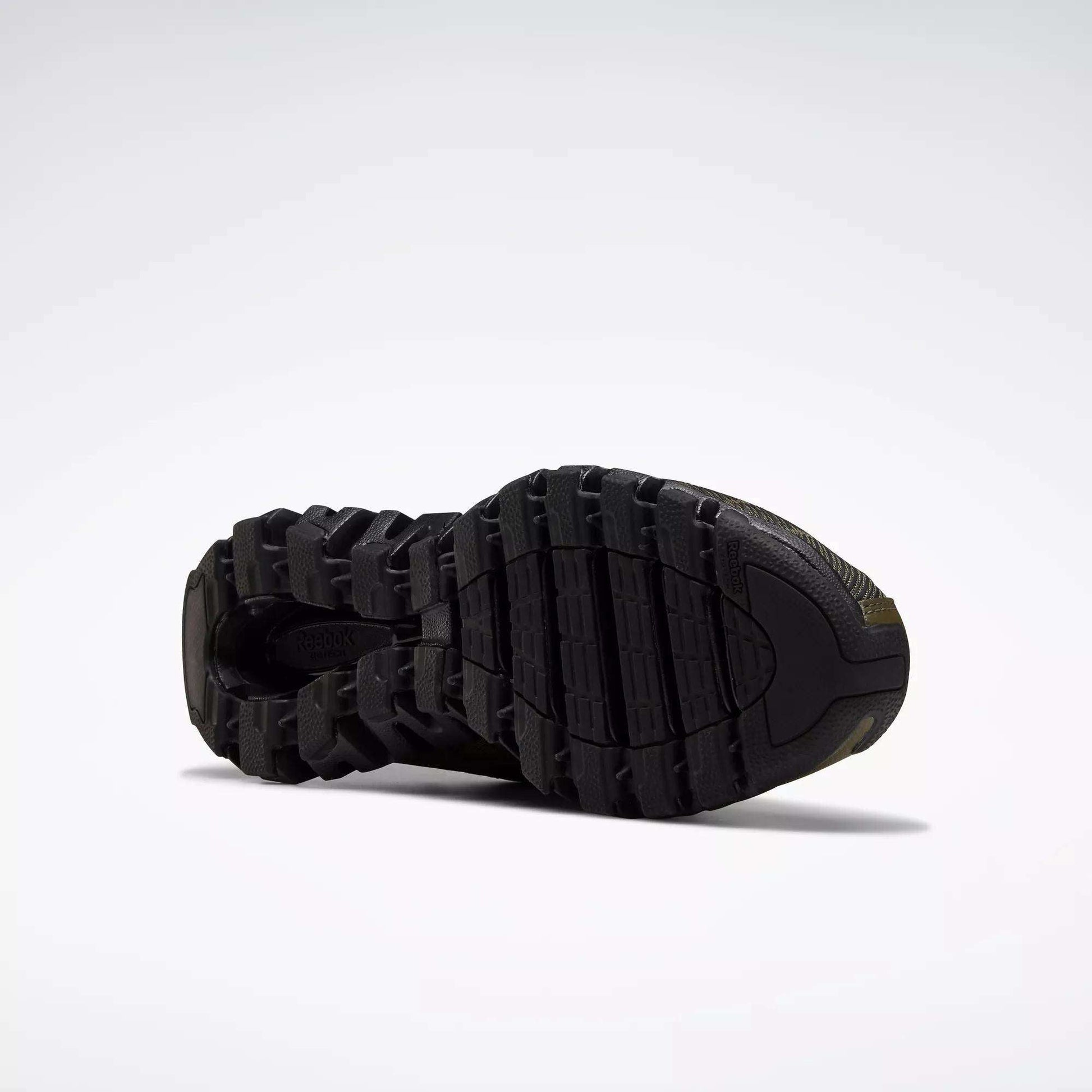 The image features the black outsole of Reebok Men's ZigWild Trail 6 Shoes, showcasing a rugged, textured tread for traction and durability, set against a plain light gray background.