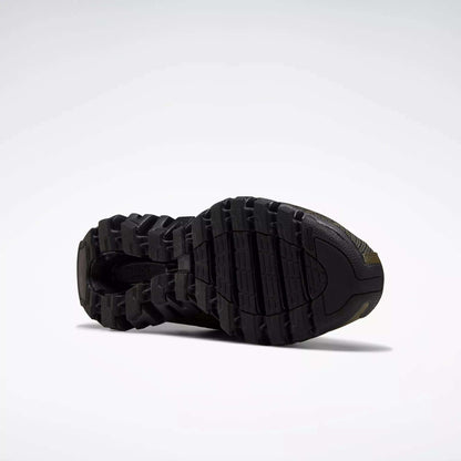 The image features the black outsole of Reebok Men's ZigWild Trail 6 Shoes, showcasing a rugged, textured tread for traction and durability, set against a plain light gray background.