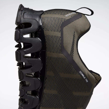 A close-up side view of the Reebok Men's ZigWild Trail 6 Shoes in black, highlighting the unique zigzag-shaped sole for cushioning and a textured upper, set against a light background.