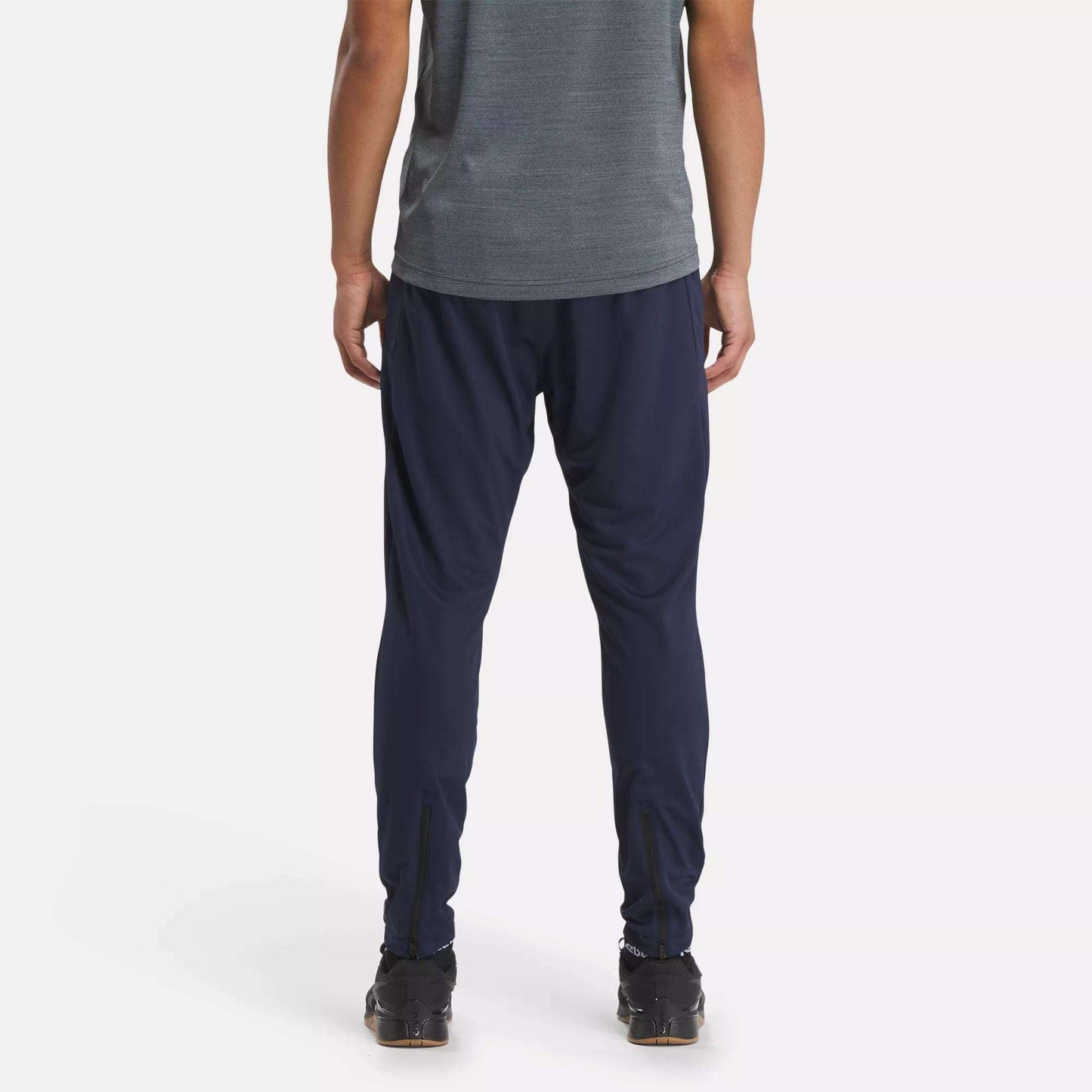 A person stands with their back to the camera, wearing a gray short-sleeve shirt and Reebok Men's Workout Ready Track Pant in navy blue, paired with black athletic shoes, against a plain white background.