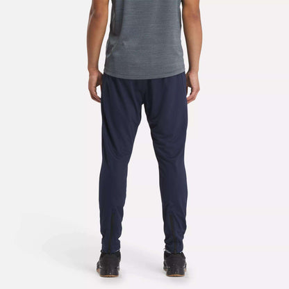 A person stands with their back to the camera, wearing a gray short-sleeve shirt and Reebok Men's Workout Ready Track Pant in navy blue, paired with black athletic shoes, against a plain white background.