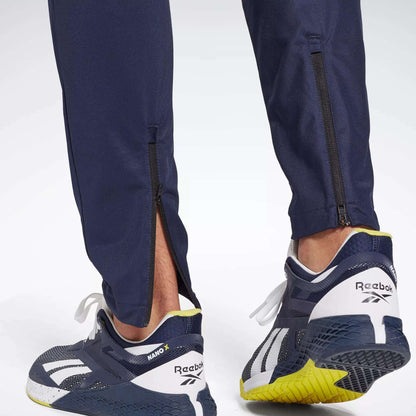 Close-up of a person in Reebok Men's Workout Ready Track Pant with ankle zippers and navy Reebok sneakers featuring white and yellow soles, focusing on the back of the shoes and lower legs.