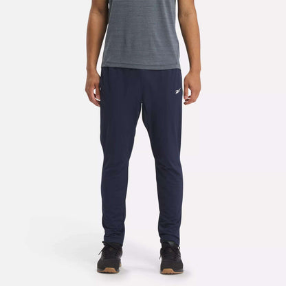 A person stands against a white background wearing Reebok Men's Workout Ready Track Pant in navy blue with a small white logo on the left thigh, paired with a grey short-sleeve shirt and black sneakers.