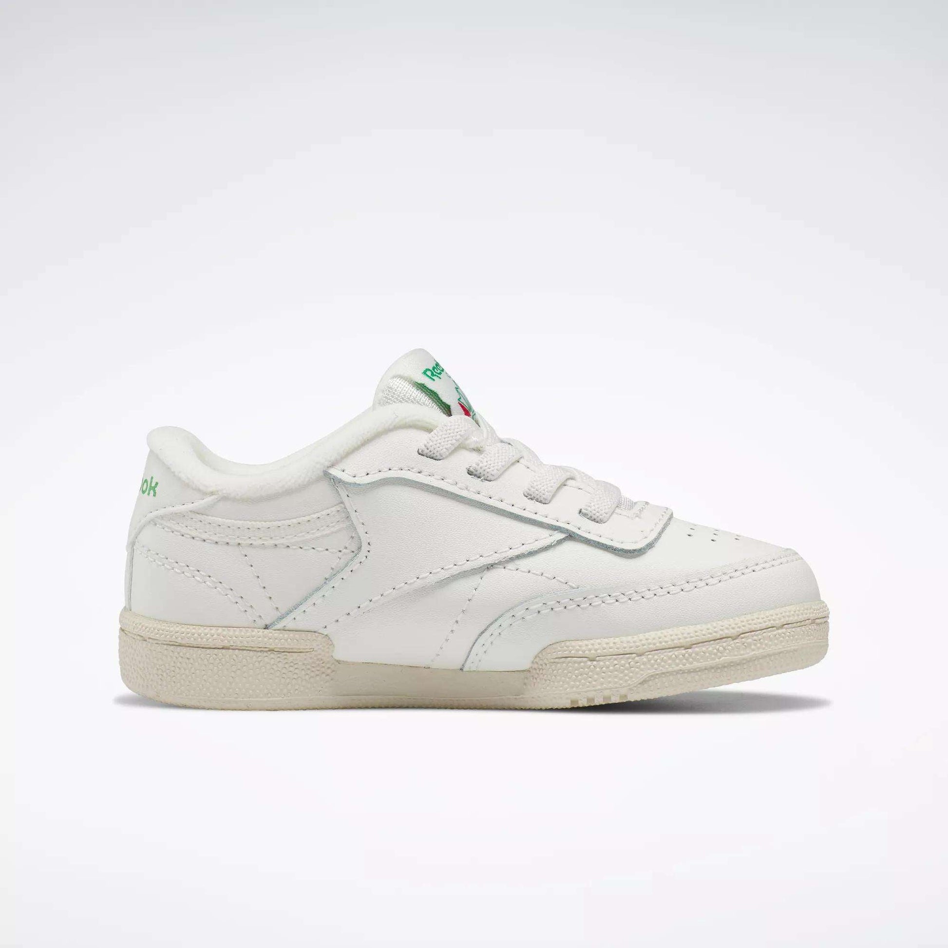 The Reebok Kids' Club C Shoes for baby & toddler feature a white design with green accents, '80s-inspired low-top style, lace-up closure, and beige rubber sole, shown on a plain light background.