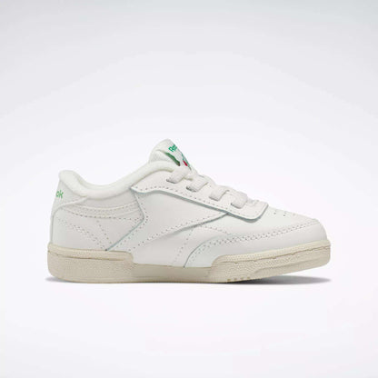 The Reebok Kids' Club C Shoes for baby & toddler feature a white design with green accents, '80s-inspired low-top style, lace-up closure, and beige rubber sole, shown on a plain light background.