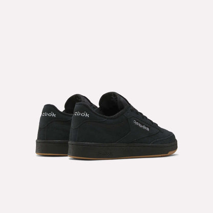 A pair of black Reebok Club C 85 Shoes with suede uppers and gum soles is displayed on a white background, one shoe facing forward and the other angled back, showcasing the court-inspired Reebok logos on the sides and heel.