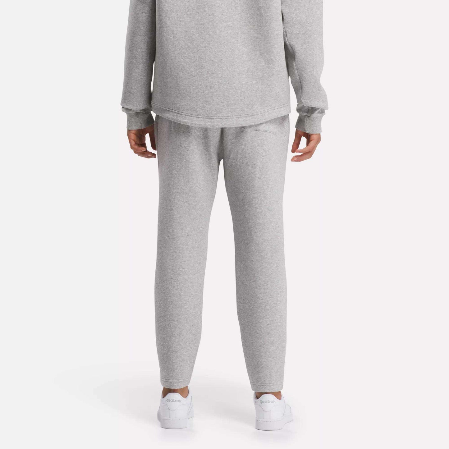 A person is seen from behind in a plain, light setting, wearing a light gray sweatshirt, white sneakers, and Reebok Men's Active Collective DreamBlend Pants with a tapered fit.