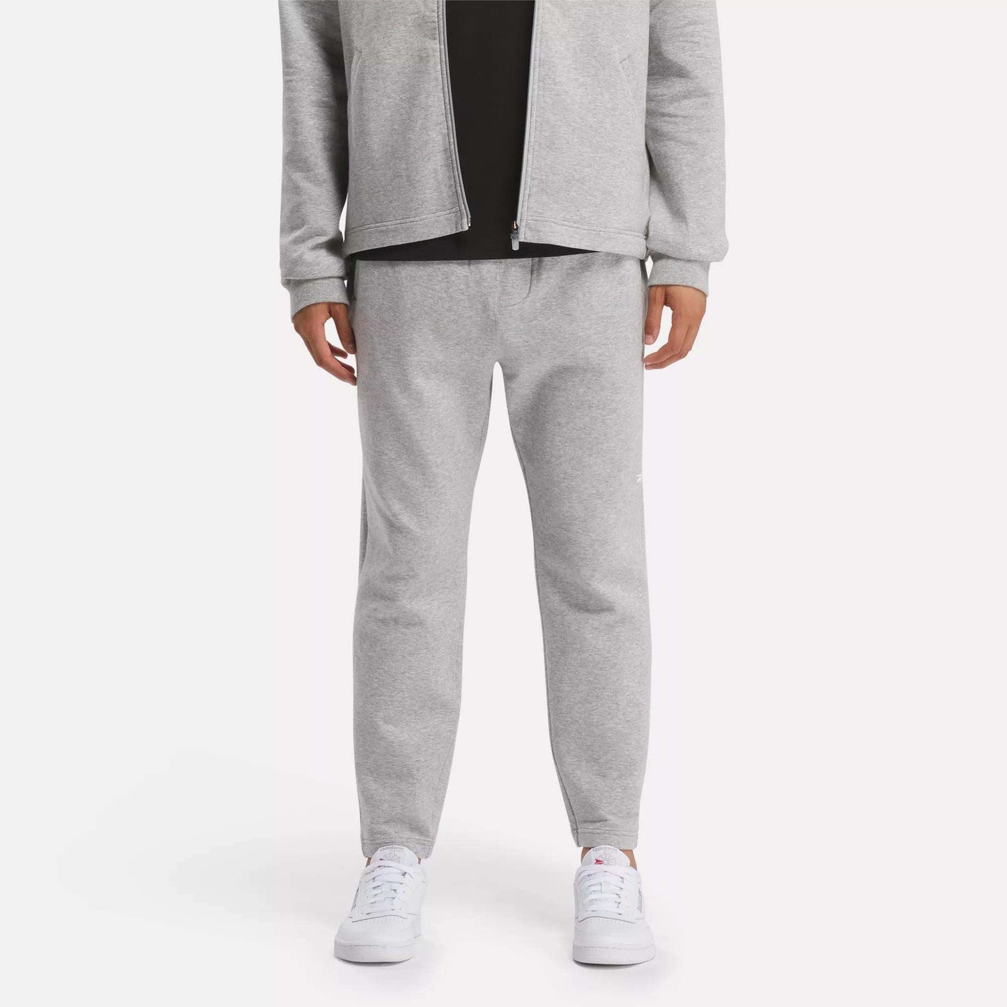 A person in a light gray zip-up jacket and Reebok Men's Active Collective DreamBlend Pants with white sneakers stands against a plain light background; their face is not visible.
