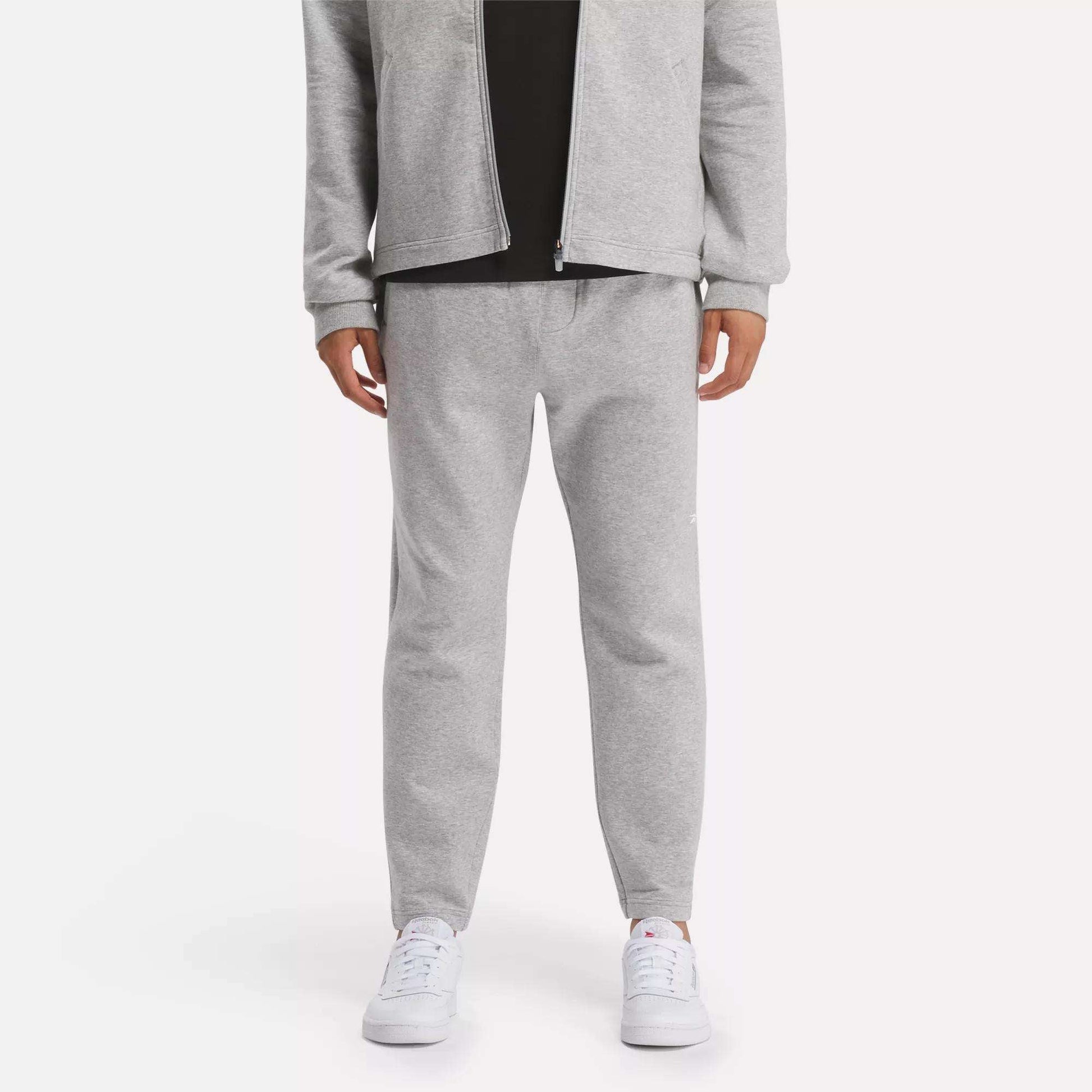 A person in a light gray zip-up jacket and Reebok Men's Active Collective DreamBlend Pants with white sneakers stands against a plain light background; their face is not visible.