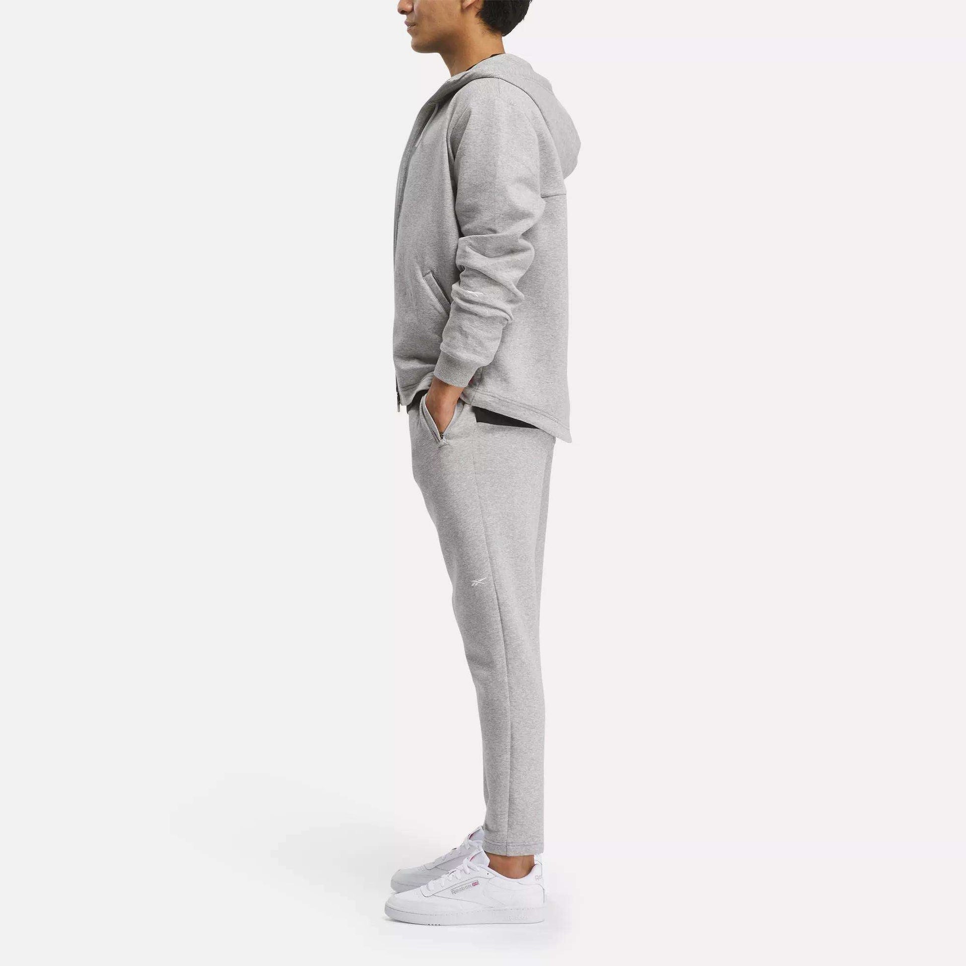 A person stands in profile, hands in pockets, wearing a light gray Reebok Men's Active Collective DreamBlend Pants with a matching hoodie and white sneakers against a plain white background.