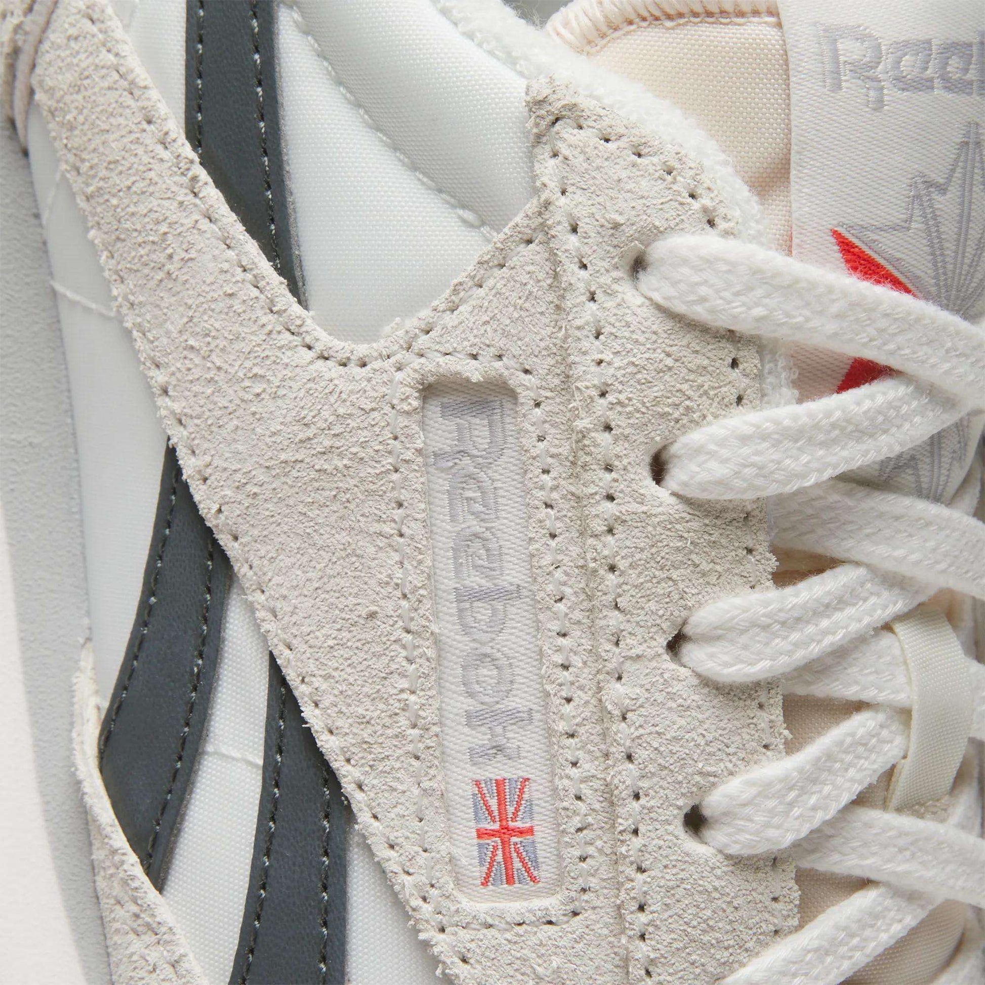 Close-up of the Women's Classic AZ Shoes by Reebok in white and beige, featuring suede and leather accents plus a woven logo tag with a small Union Jack near the laces—ideal for vintage sneaker enthusiasts.